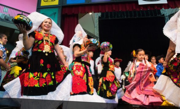 Several people dance at a community celebration, wearing brightly colored dresses and carrying art.