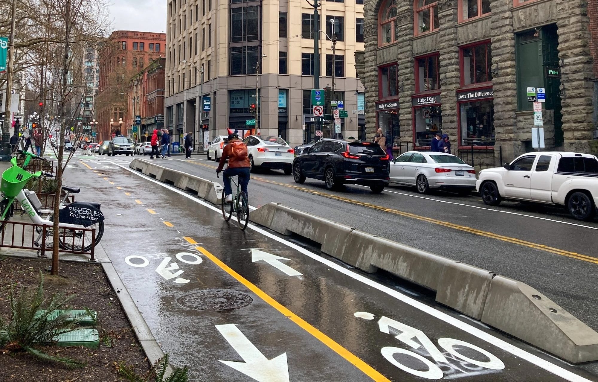 A person bikes up a protected bike lane on a damp day with buildings and cars in the background.