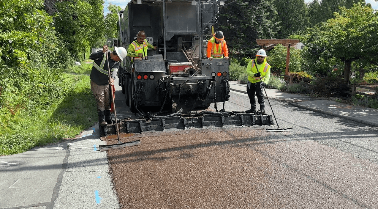 Four workers wearing high visibility vests work to apply street sealant using a large vehicle. Trees and plants are in the background.