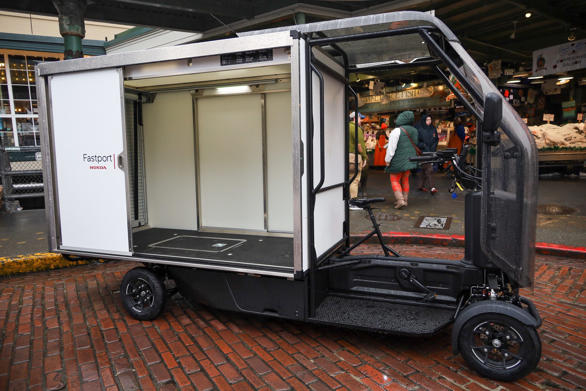 A large commercial e-bike with storage in the back is parked on brick cobblestones at a public market. People are in the background.