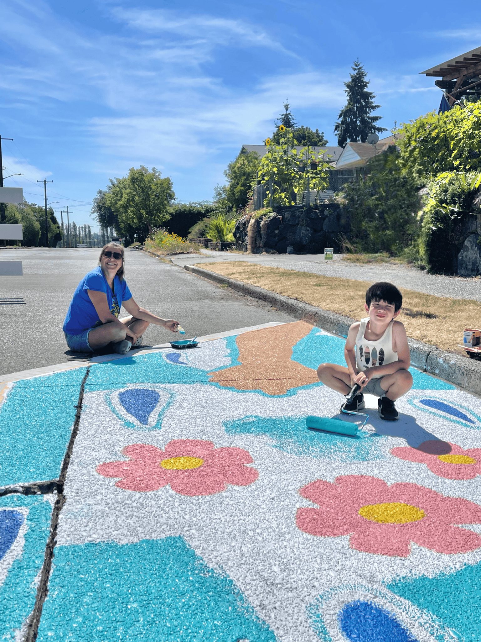 Two people including a child smile while working on a large, colorful mural painted on the street, with houses and trees in the background.