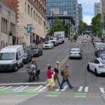 Several people walk across the street at a marked crosswalk in the city on a clear day. Cars and a motorcycle are in the background, as well as several parked cars and large buildings.