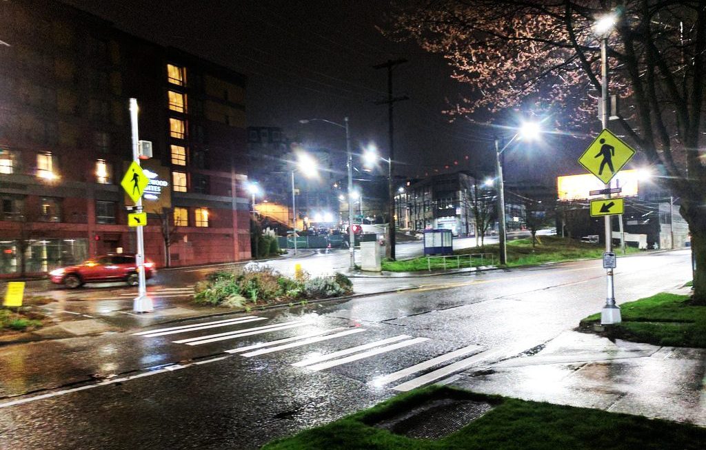 A marked crosswalk with flashing signals or beacons at night. A car drives past a large building in the background.