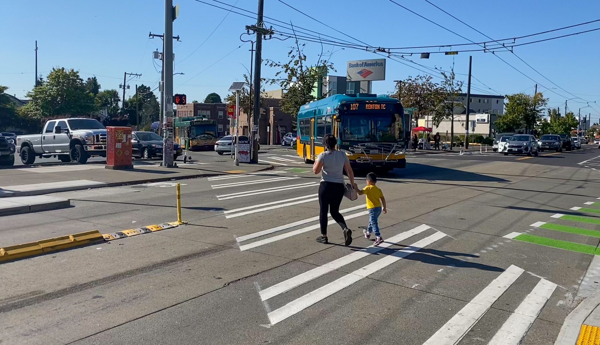 Two people walk across the street at a marked crosswalk in the city on a busy day. A bus prepares to turn onto the street from a nearby street.