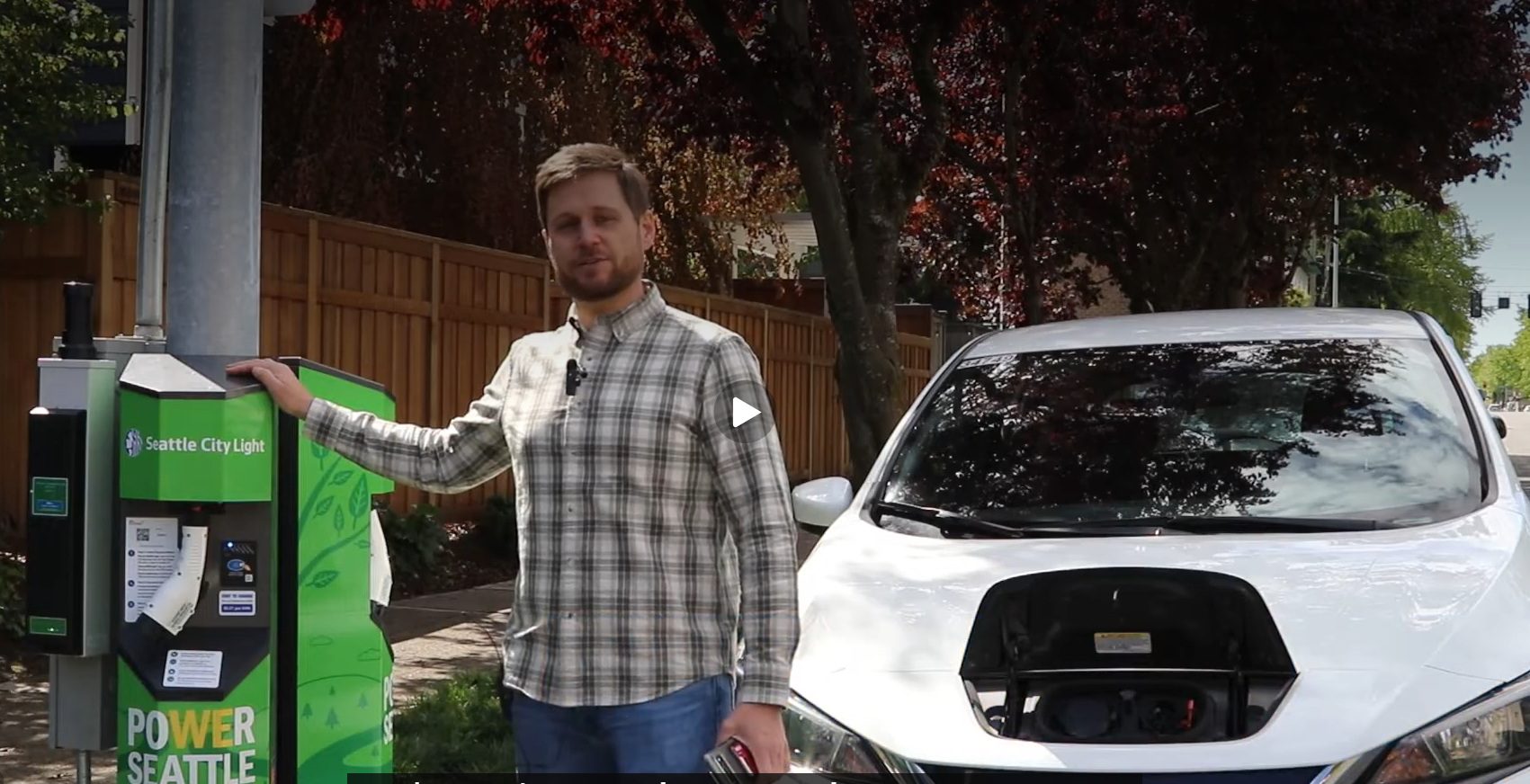 A man stands next to a green electric vehicle charging station with a white car to the right, and trees and a fence in the background on a clear day.