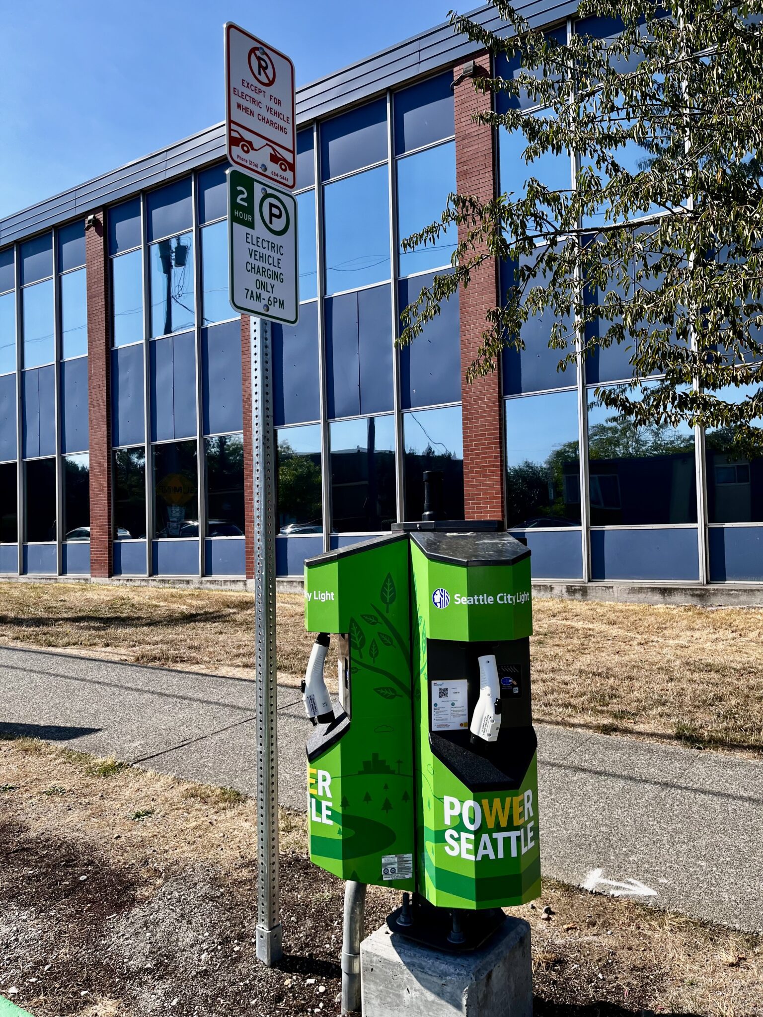 A bright green electric vehicle charging station sits at the curb in front of a large building on a clear, sunny day.