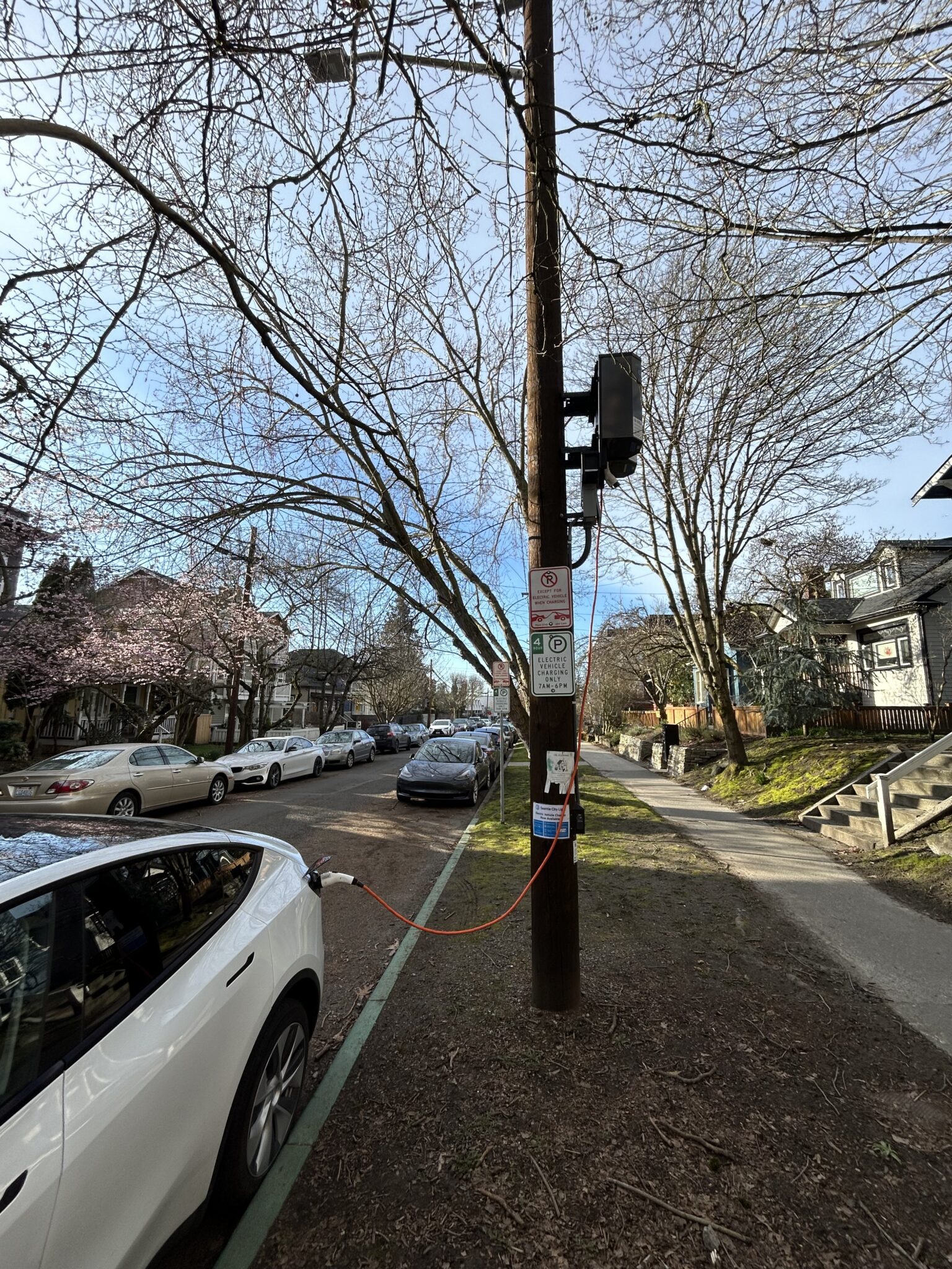 A white electric vehicle is charging at a curbside parking spot on a clear sunny day. Parked cars, trees, and houses are in the background.